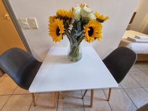 A small table with a vase of sunflowers and a white tablecloth, flanked by two grey chairs in a room with a bed in the background.