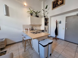 A compact kitchen with a central island, bar stools, a sink, and a stove. There are potted plants on the island and a coat rack to the right.