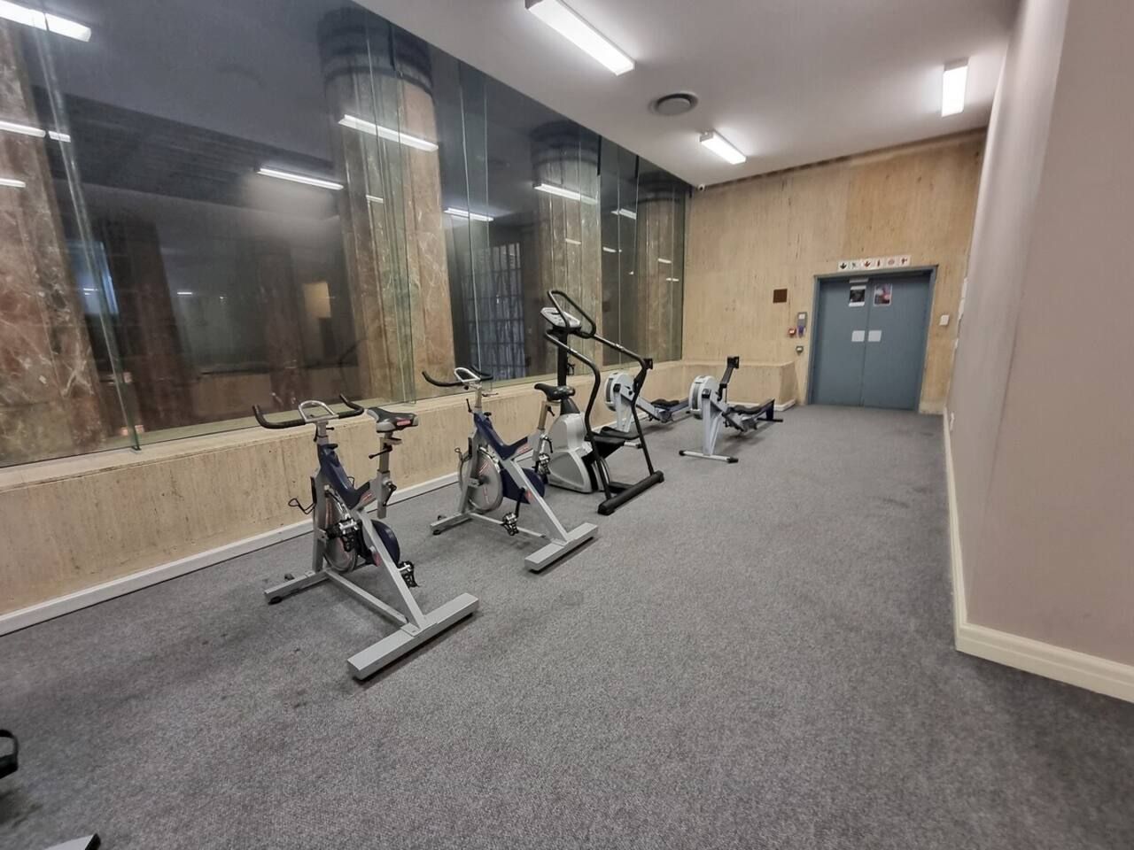 Une salle de sport avec des vélos d’appartement alignés en rangée, une porte d’ascenseur bleue et une colonne en marbre.