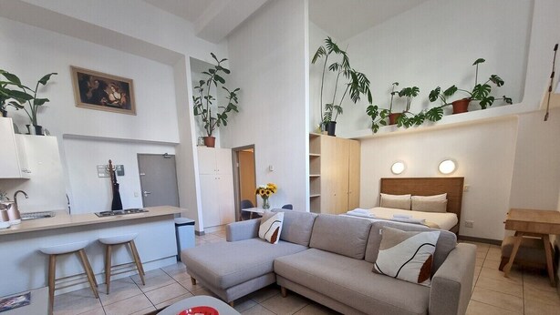 A modern living room with a grey sofa, a dining table with two stools, a kitchen area with a fridge, and a bed with a headboard.