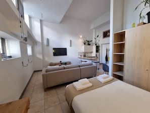 Una camera moderna e luminosa con un divano, un letto, una TV e un angolo cottura.