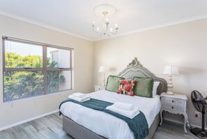 A bedroom with a bed, two bedside tables, a nightstand, a chandelier, and a window with blinds.