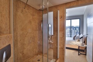 Casa de banho moderna com cabine de duche envidraçada, lavatório visível e cama com mesa de cabeceira no quarto adjacente.