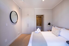 A hotel room with a bed, a mirror, a wardrobe, a bathroom with a shower, and a nightstand with a lamp.