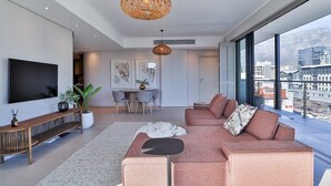 Un salon moderne doté d’un grand téléviseur à écran plat, d’une console en bois, d’un grand canapé avec des coussins, d’un coin repas avec une table et des chaises, et d’une grande fenêtre offrant une vue sur la ville.