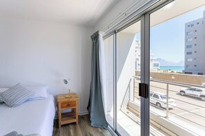 A hotel room with a bed, a nightstand, a lamp, and a large window with a view of a cityscape and vehicles. - 2br Coastal Stay | Walk to beach + bbq by ctha (Cape Town)