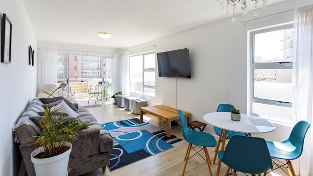A modern living room with a sofa, a coffee table, and a dining area with chairs. There is a TV mounted on the wall and a plant on the sofa.