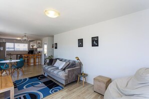 A modern living room with a grey sofa, a coffee table, and a dining area with chairs. - 2br Coastal Stay | Walk to beach + bbq by ctha (Cape Town)