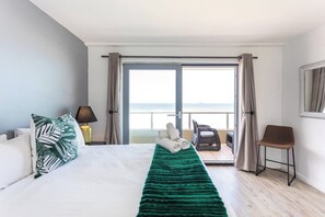 Una camera d'albergo con un letto, una coperta verde, una sedia, una lampada e vista sul mare attraverso un'ampia finestra.