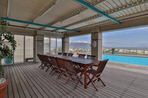 Una terrazza esterna con tavolo e sedie in legno, affacciata su una piscina e una spiaggia.