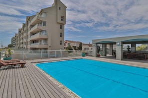 Una piscina con un pontile in legno, un'area salotto coperta e un edificio a più piani sullo sfondo.