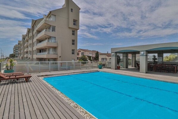 Une piscine avec une terrasse en bois, un espace salon couvert et un immeuble à plusieurs étages en arrière-plan.
