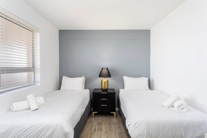 A hotel room with two beds, a nightstand with a lamp, and a window with blinds.