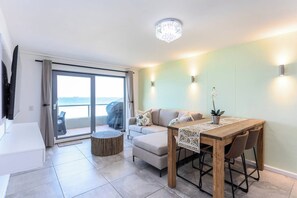 A modern living room with a sofa, a dining table, and a view of the sea through large sliding doors.