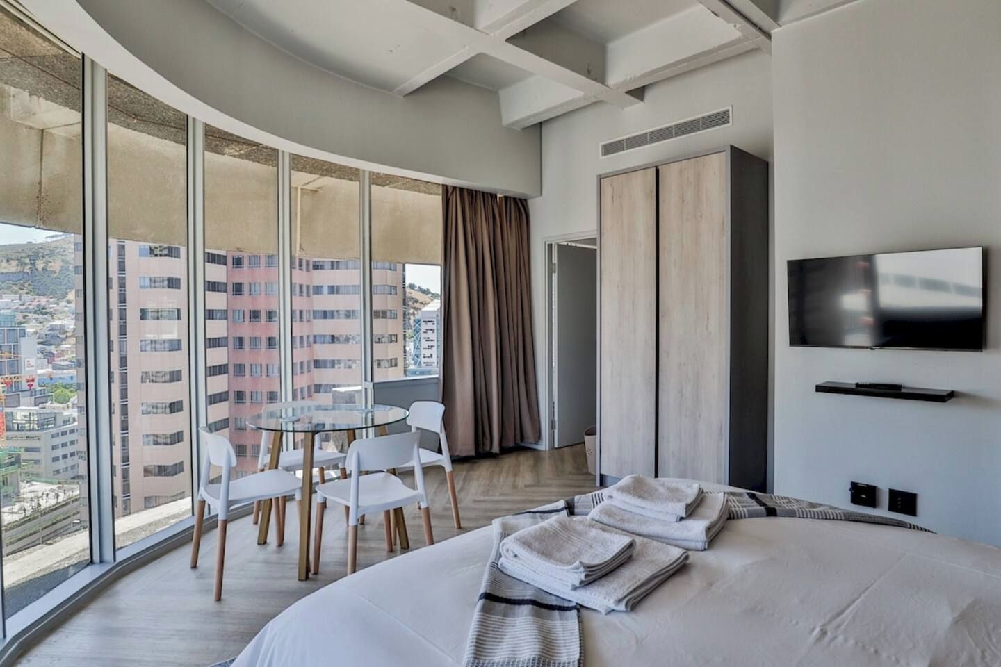 Quarto de hotel com uma cama grande, televisão, mesa de jantar com cadeiras e vista da cidade através de grandes janelas.
