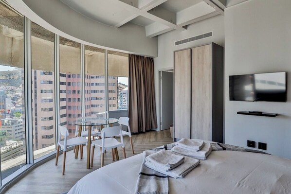 A hotel room with a large bed, a TV, a dining table with chairs, and a view of the city through large windows.