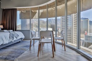 Una camera d'albergo moderna con un letto grande, un tavolo da pranzo con sedie e vista sulla città attraverso finestre a tutta altezza.