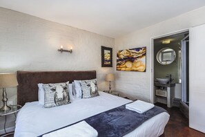 A hotel room with a large bed, a headboard, a wall-mounted light fixture, a painting, and a bathroom visible through an open door.