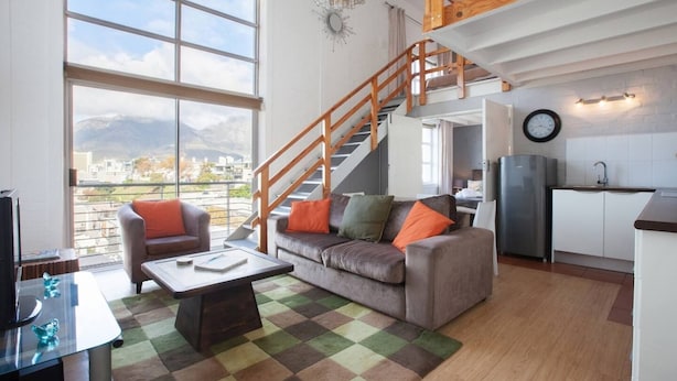 A modern living space with a sofa, armchair, and coffee table, a staircase, and a kitchen area. - Harbour Terrace 48 by Ctha (Cape Town)