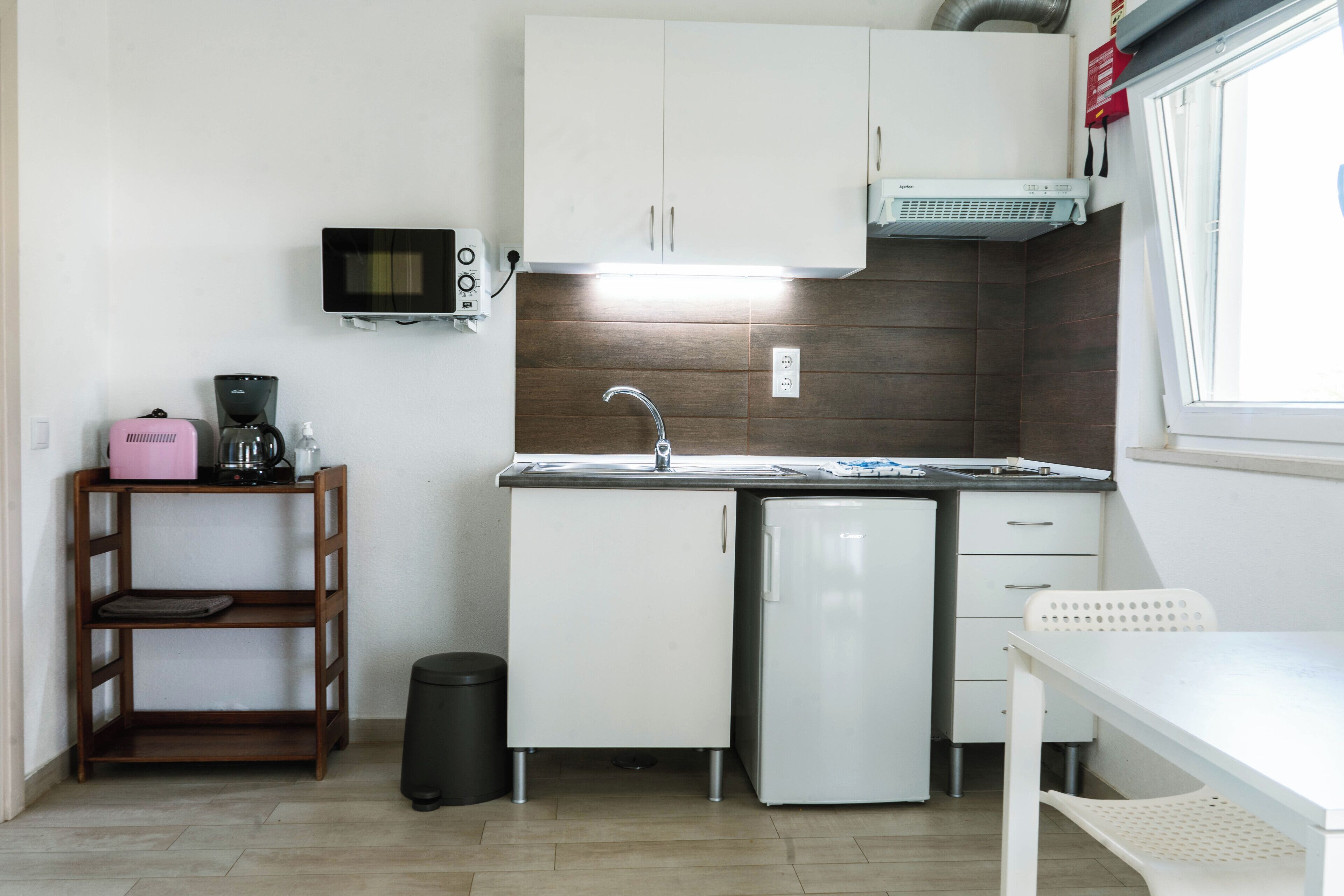 Eine kompakte Küchenzeile mit Mikrowelle, Spüle und kleinem Kühlschrank. Ein Holzregal beinhaltet einen Wasserkocher und einen Mülleimer.