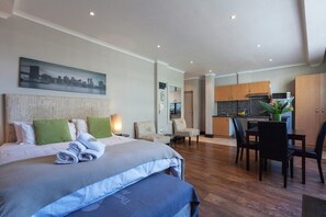 A hotel room with a bed, a dining table, chairs, a kitchen area, and a cityscape painting on the wall.