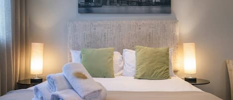 A hotel room with a bed, two pillows, two bedside lamps, and a cityscape painting above the bed.