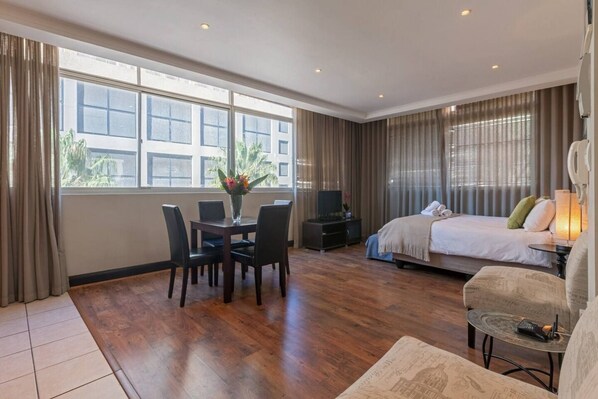 A hotel room with a bed, a sofa, a dining table with chairs, a TV, and a window with curtains.