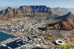 An aerial view of a coastal city with a prominent mountain range in the background, a marina with boats, and a large stadium.
