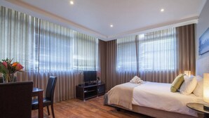 A hotel room with a bed, a desk with a chair, a TV, and curtains. - City Studio | Parking, Pool & gym by ctha (Cape Town)