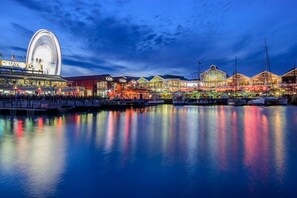 A waterfront area with a Ferris wheel, various shops, and boats docked along the water.