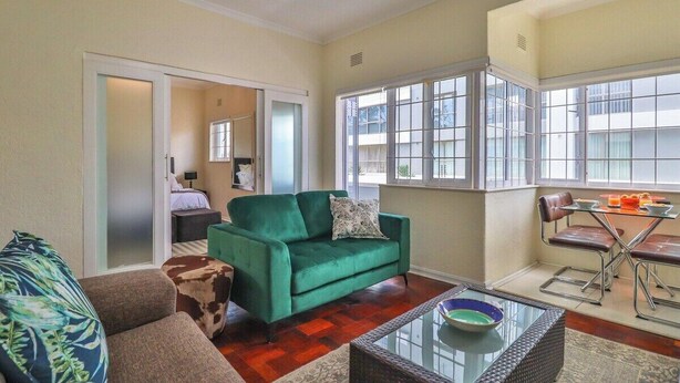 A living room with a green sofa, a brown sofa, a glass coffee table, and a dining area with a metal table and chairs. - Eaton Square 2 by CTHA (Cape Town)