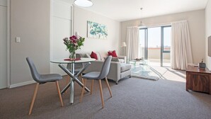 Een moderne woonkamer met een glazen eettafel, twee grijze stoelen, een bank met rode kussens, een schilderij aan de muur en uitzicht door een groot raam.