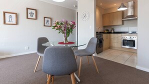 Un apartamento moderno con comedor que incluye una mesa de vidrio y sillas grises, cocina con electrodomésticos de acero inoxidable, y un reloj de pared.