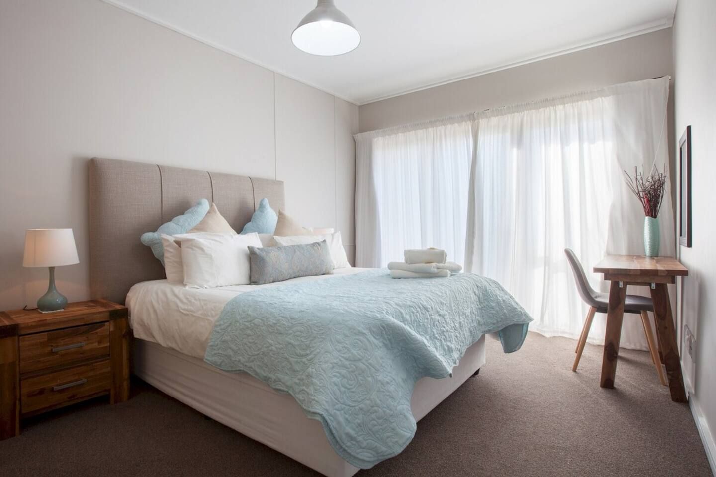 A bedroom with a bed, bedside table, lamp, and a desk with a chair.