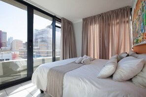 Een hotelkamer met een groot bed, uitzicht op de stad door de vloerhoogte ramen en een balkon met een hekwerk.