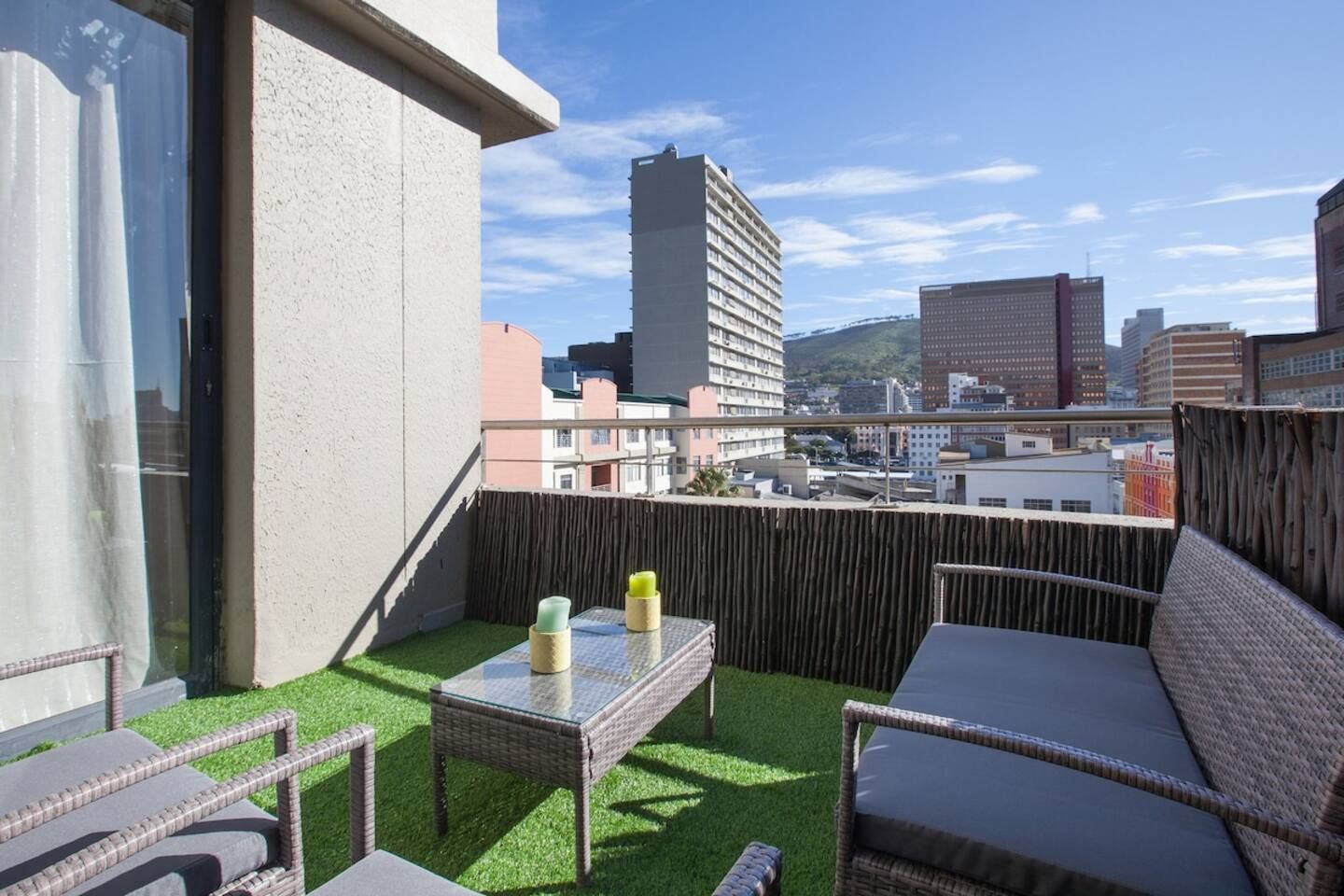 A balcony with a wicker table and chairs, a view of the cityscape, and a wooden fence.