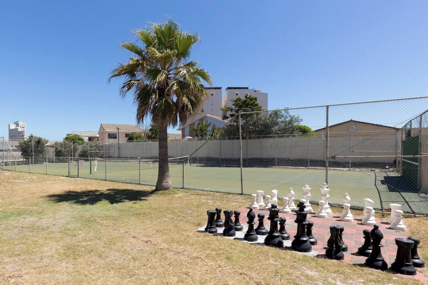 Ein eingezäunter Tennisplatz mit einer Palme und Schachfiguren im Vordergrund.