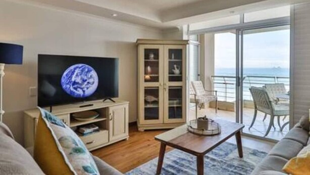 A living room with a television, a cabinet, a sofa, a table, and a balcony with a view of the sea. - Ocean View C403 by Ctha (Cape Town)