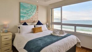 A bedroom with a bed, a nightstand, a painting, and a view of the ocean. - Ocean View C403 by Ctha (Cape Town)