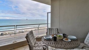 A balcony with a view of the beach, a round table with plates and a bowl, and two chairs. - Ocean View C403 by Ctha (Cape Town)