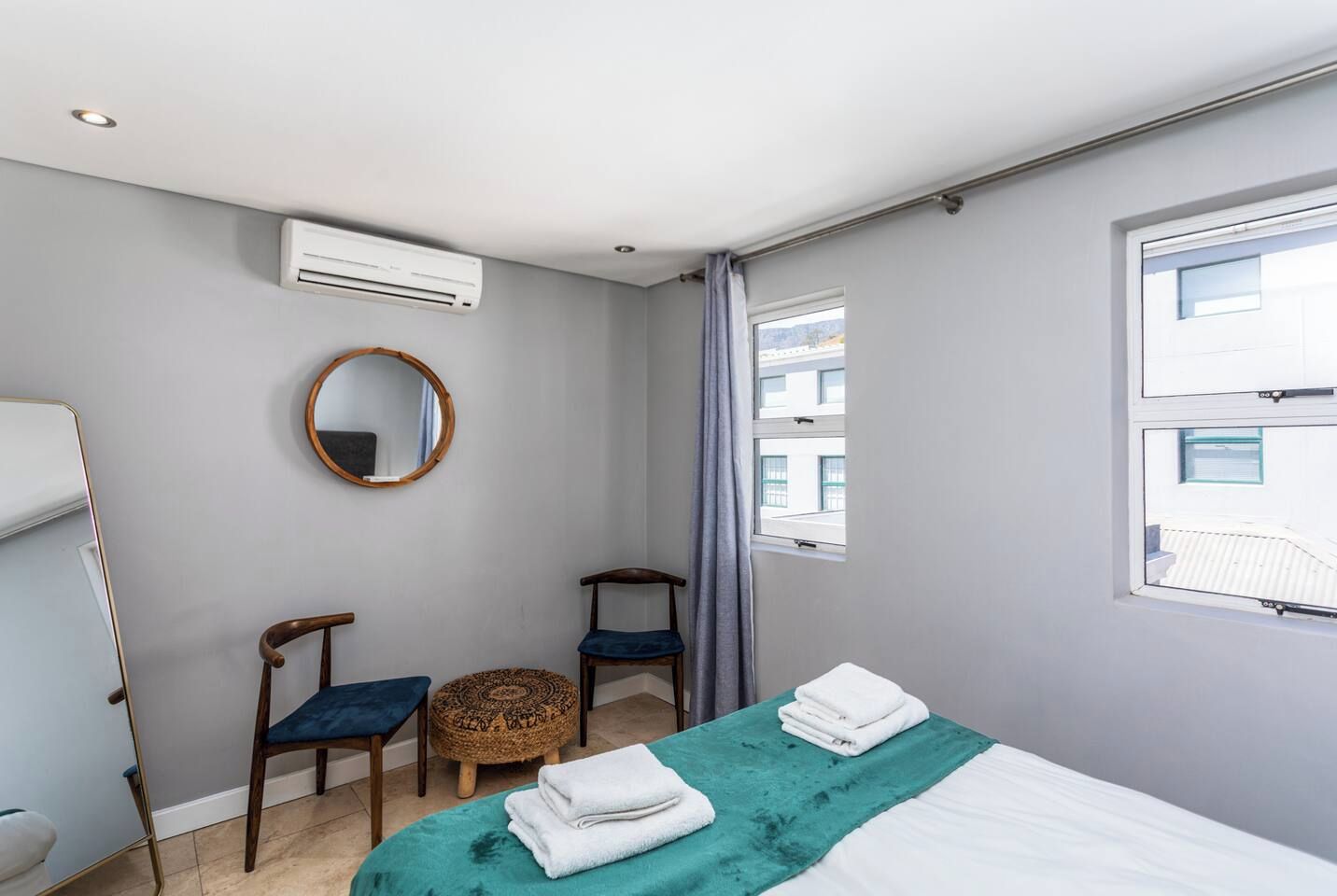 Une chambre à coucher comprenant un lit, deux chaises, un miroir rond, un appareil de climatisation et une fenêtre.