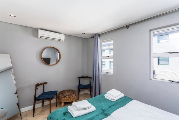 A bedroom with a bed, two chairs, a round mirror, an air conditioning unit, and a window.