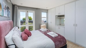 Un dormitorio con cama, una silla y un armario. Se ve una ventana con vistas a edificios y árboles.
