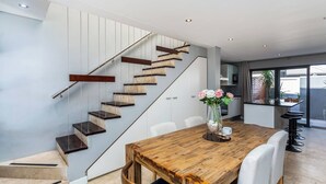 Un interior moderno con una escalera de madera, un comedor con mesa de madera y una cocina visible a través de una puerta abierta.
