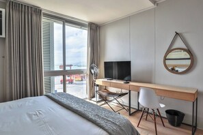 A hotel room with a bed, a desk, a chair, a television, a mirror, and a window with a view of a dock and a ship.