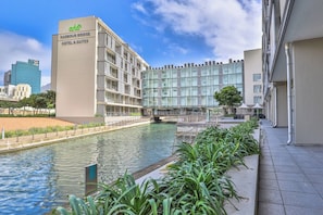 Un hotel moderno con facciata in vetro, vista sul lungomare e un ponte che lo collega a un altro edificio.