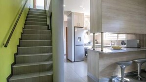 A modern kitchen with a central island, stainless steel bar stools, and a staircase leading to another level. - Modern 3 Bedroom with spacious braai area by Ctha (Century City)