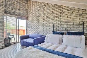 Una camera da letto con parete in mattoni, un letto con biancheria bianca, un divano blu e una porta scorrevole in vetro che dà su una zona esterna.