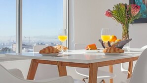 Une table dressée pour le petit-déjeuner, avec des croissants, du jus d’orange et un panier de fruits, le tout placé devant une fenêtre offrant une vue sur la ville.