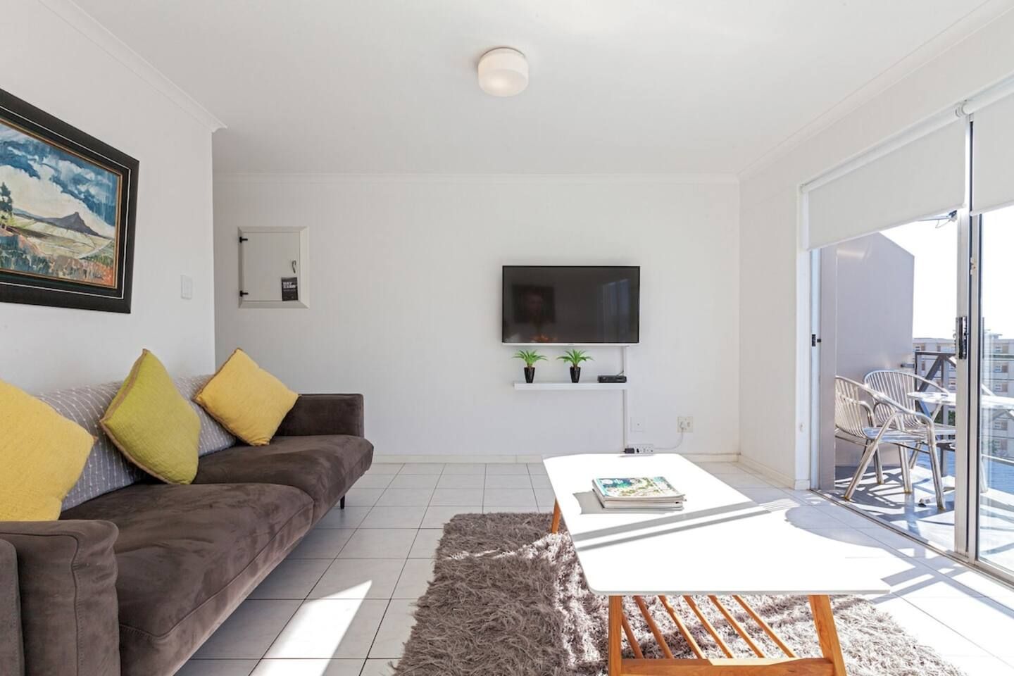 A modern living room with a sofa, a coffee table, a flat-screen TV, and a balcony with outdoor seating.